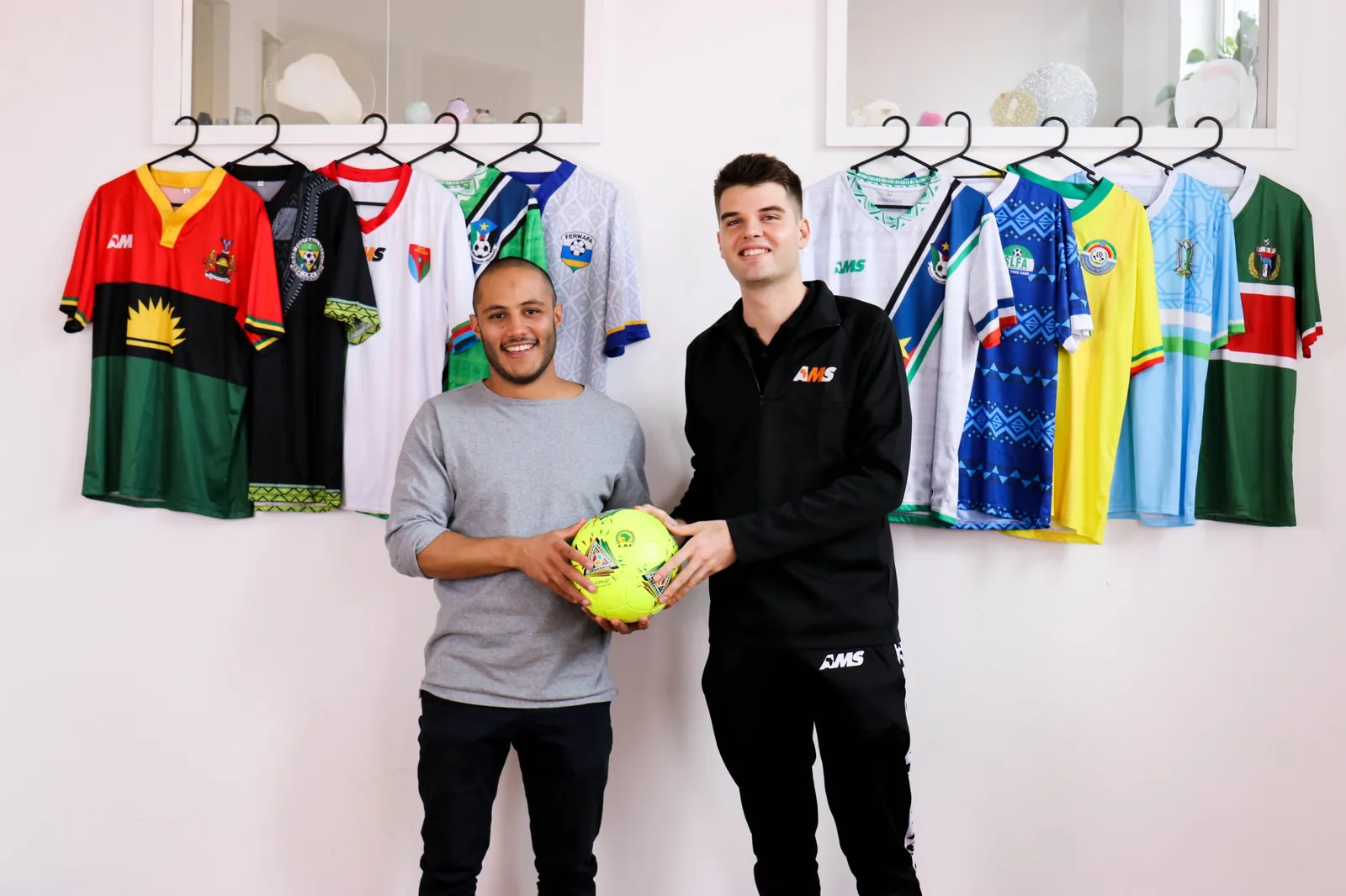 Two members of the AMS team hold a ball in front of a wall of national team shirts they designed