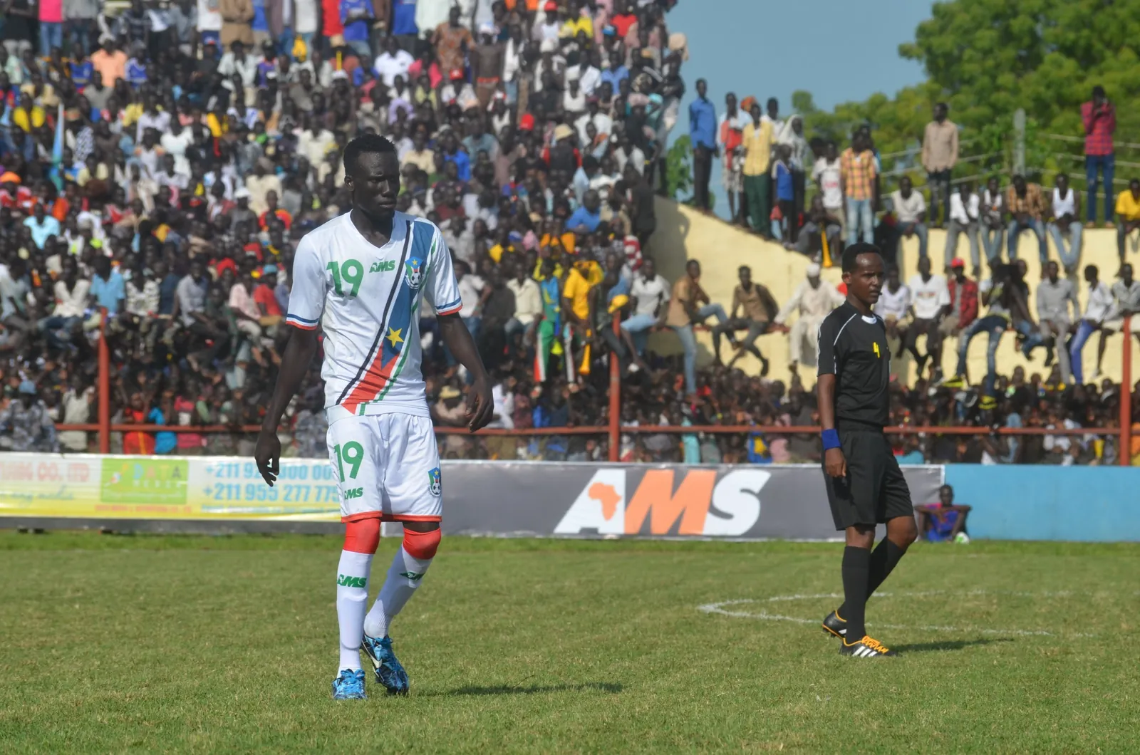 A South Sudan player in the white AMS kit with its flag-coloured sash, in front of a packed stand and an AMS advertising board