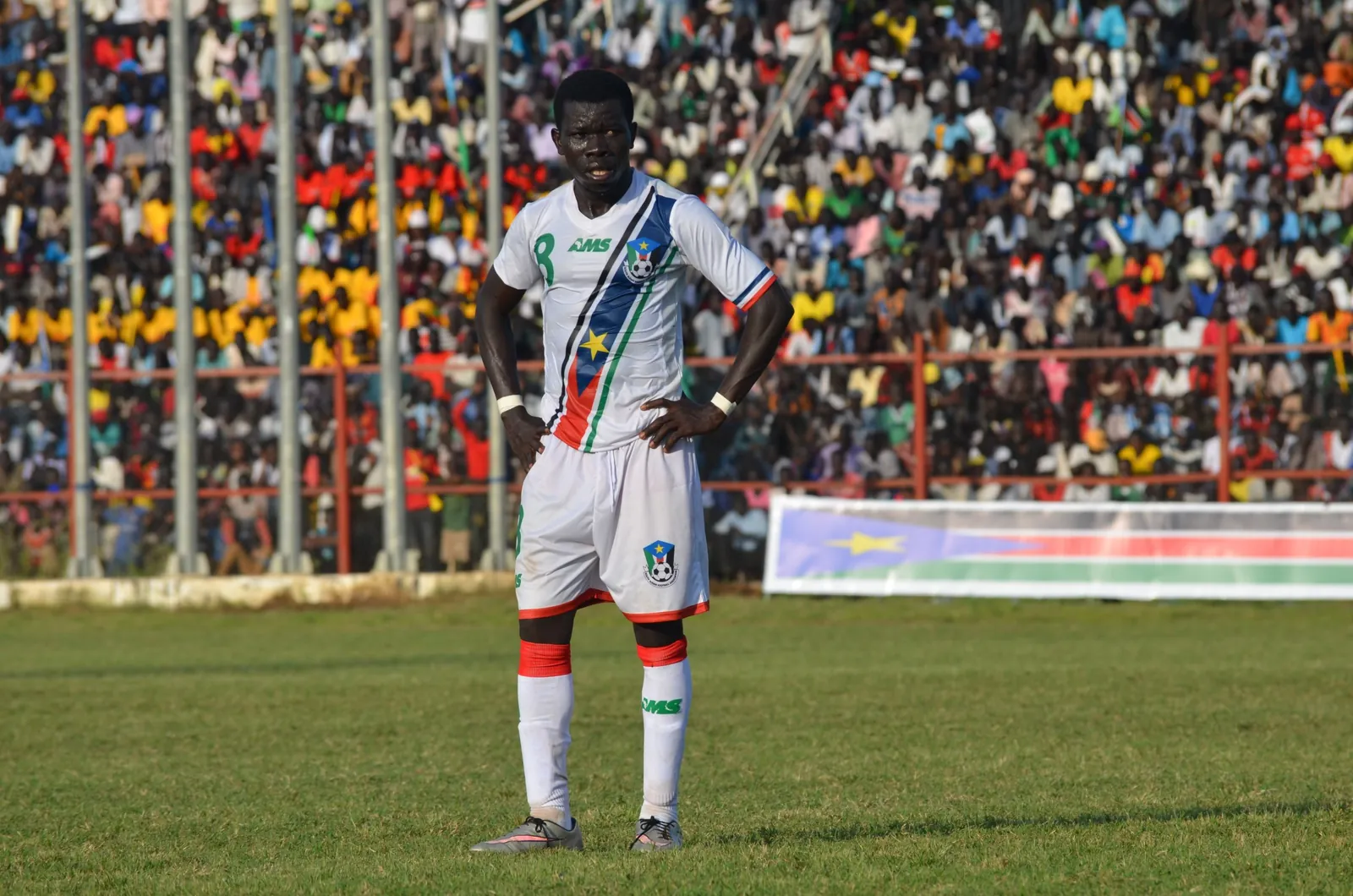 A South Sudan player stands with his hands on his hips in the white AMS kit, a full stadium behind him