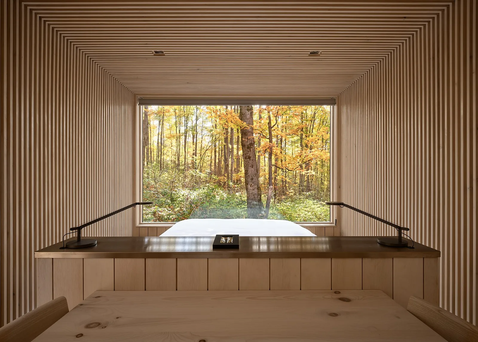 Slatted wood cabin interior with a bed below a large window framing autumn forest
