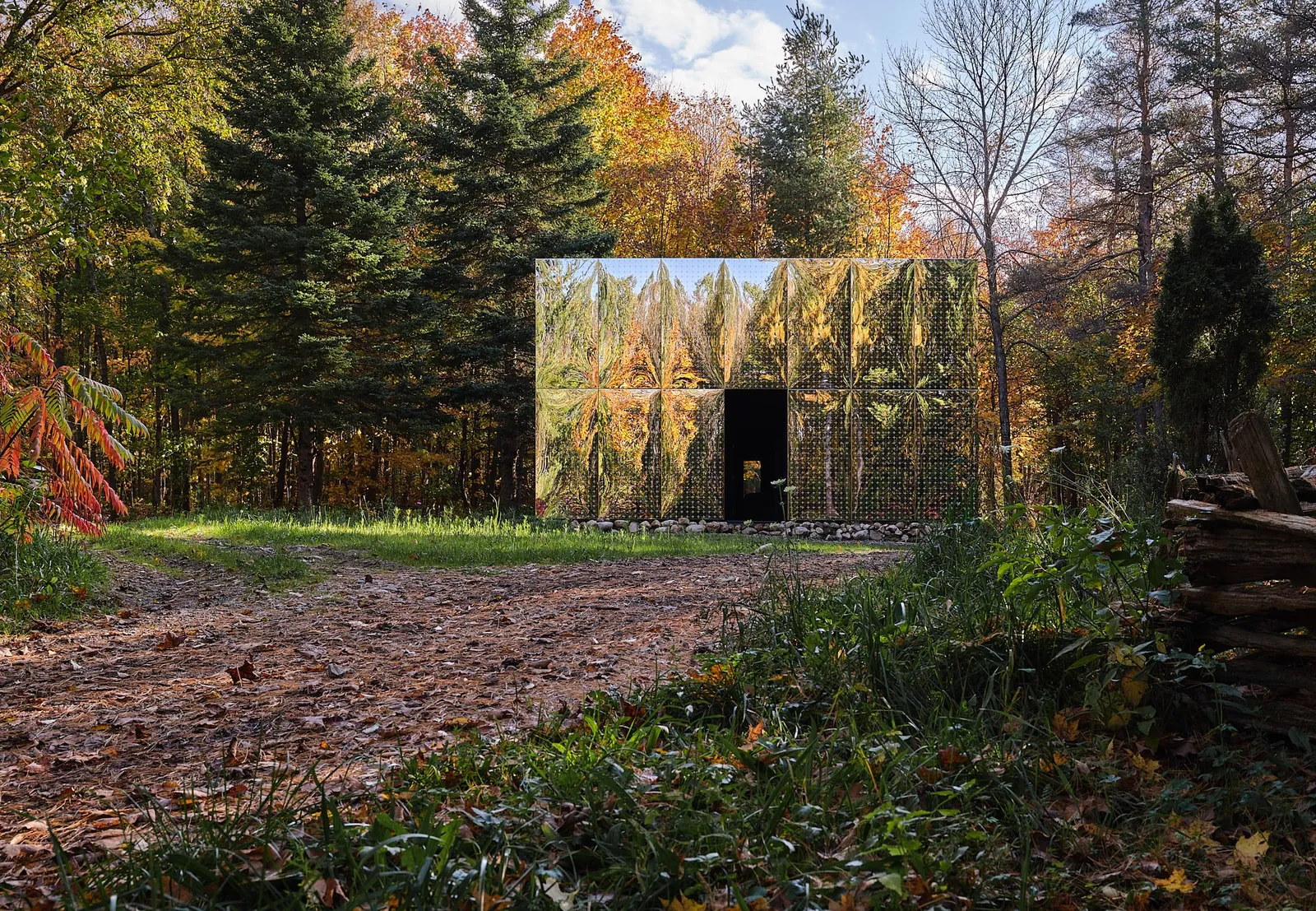 Mirrored cabin with an open doorway reflecting evergreens and autumn trees beside a forest path
