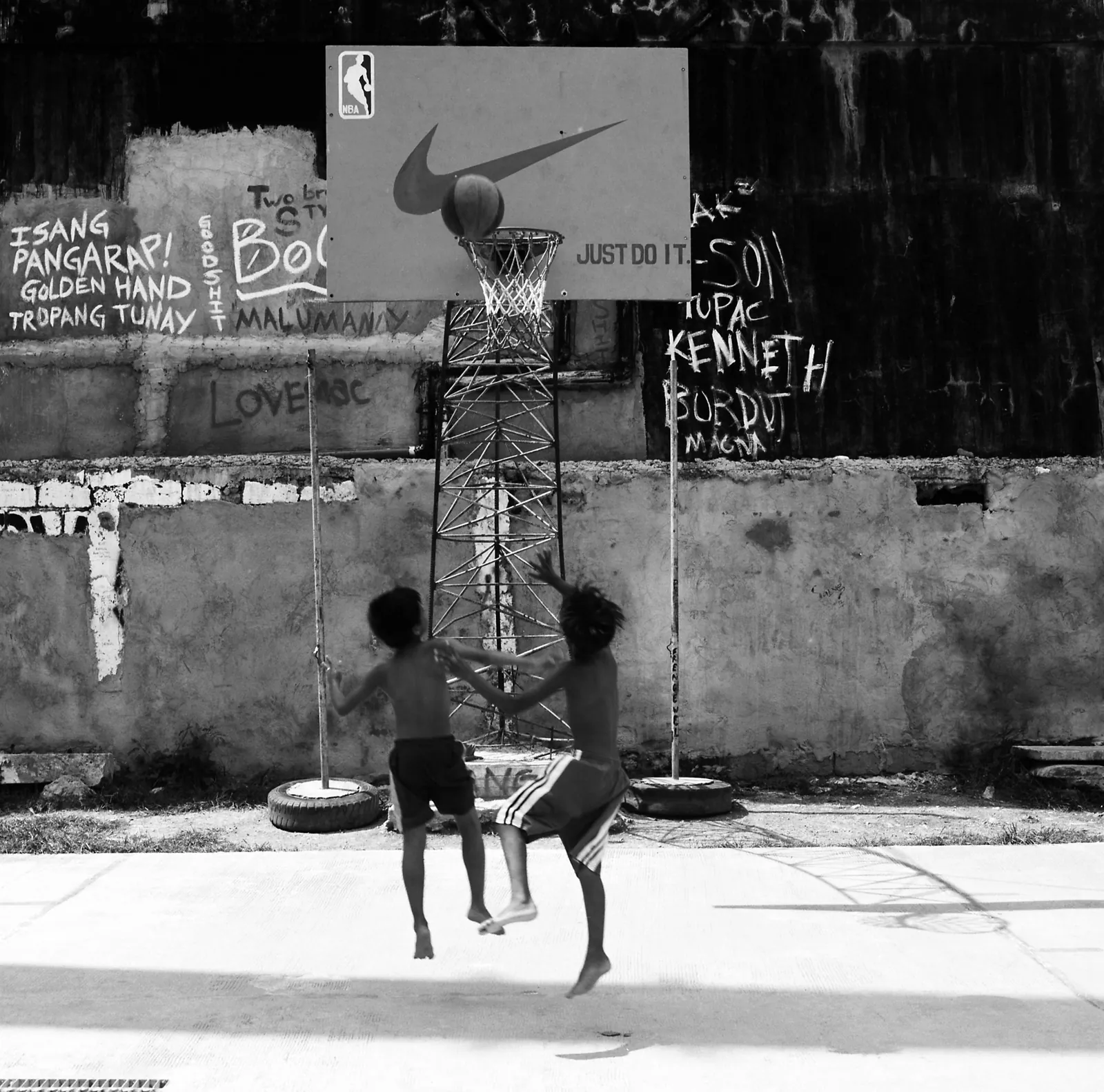 Black and white shot of two boys leaping for a ball at a Nike-logo backboard, graffiti-covered wall behind