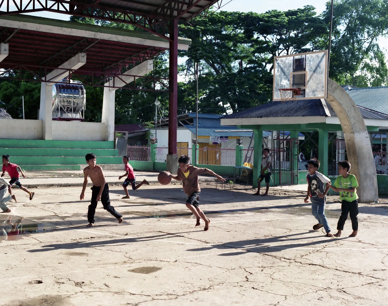 Barefoot boys playing basketball on a cracked concrete court with a covered stand and trees behind