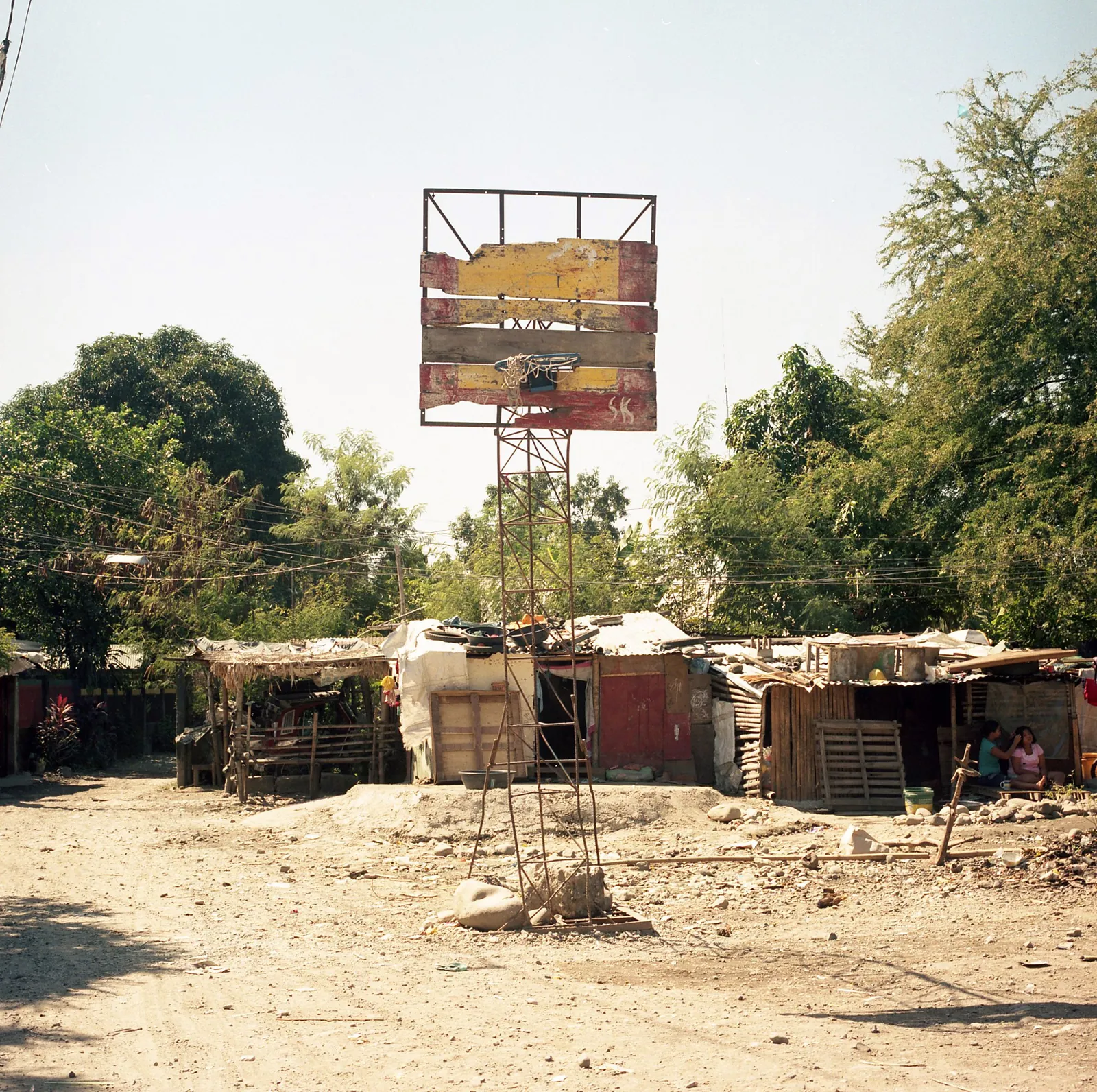 Hoop with a backboard of painted planks on a rusted metal tower, on a dirt lot beside makeshift shacks