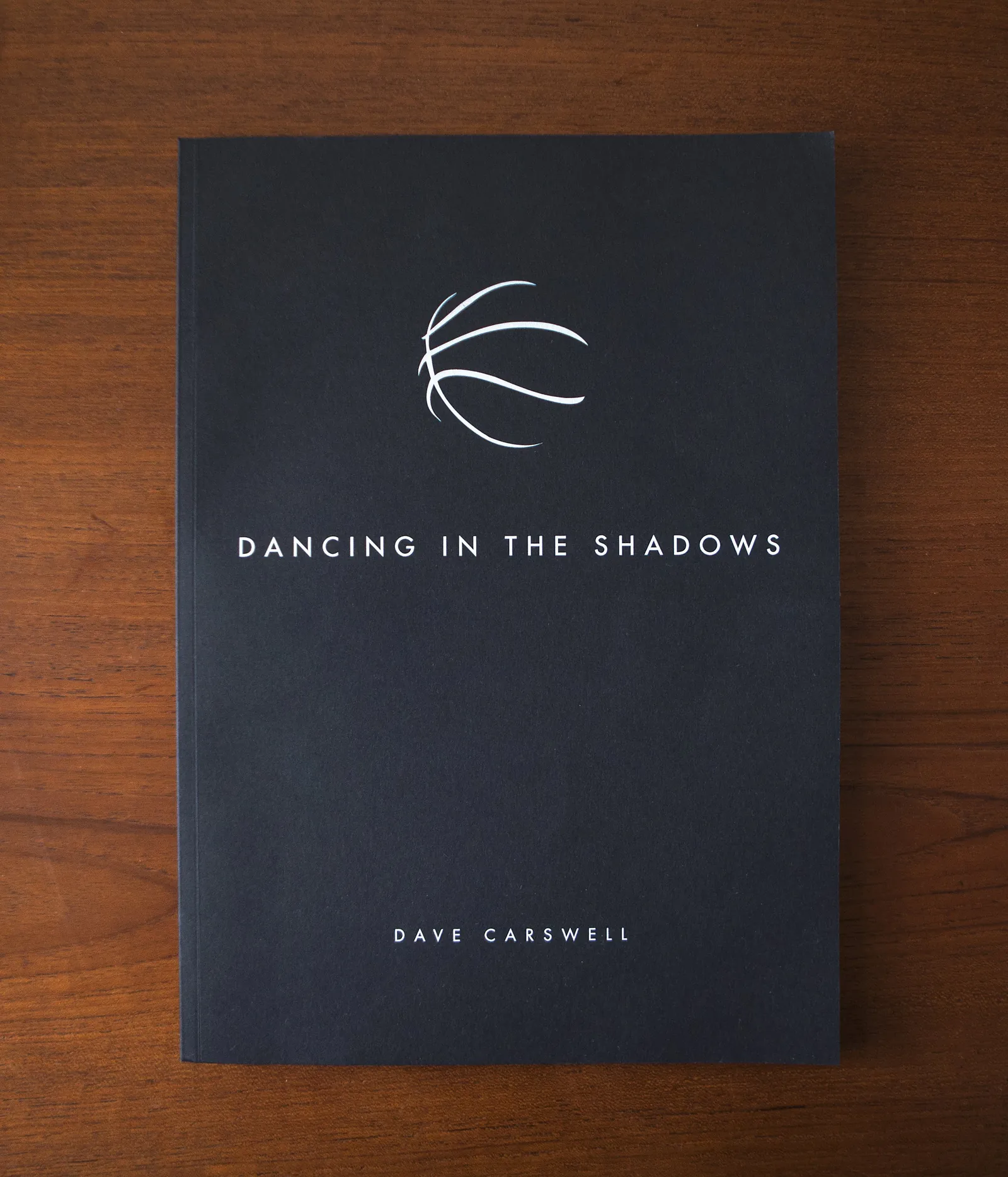 Cover of Dave Carswell’s book Dancing in the Shadows, a white basketball outline on black, on a wooden table