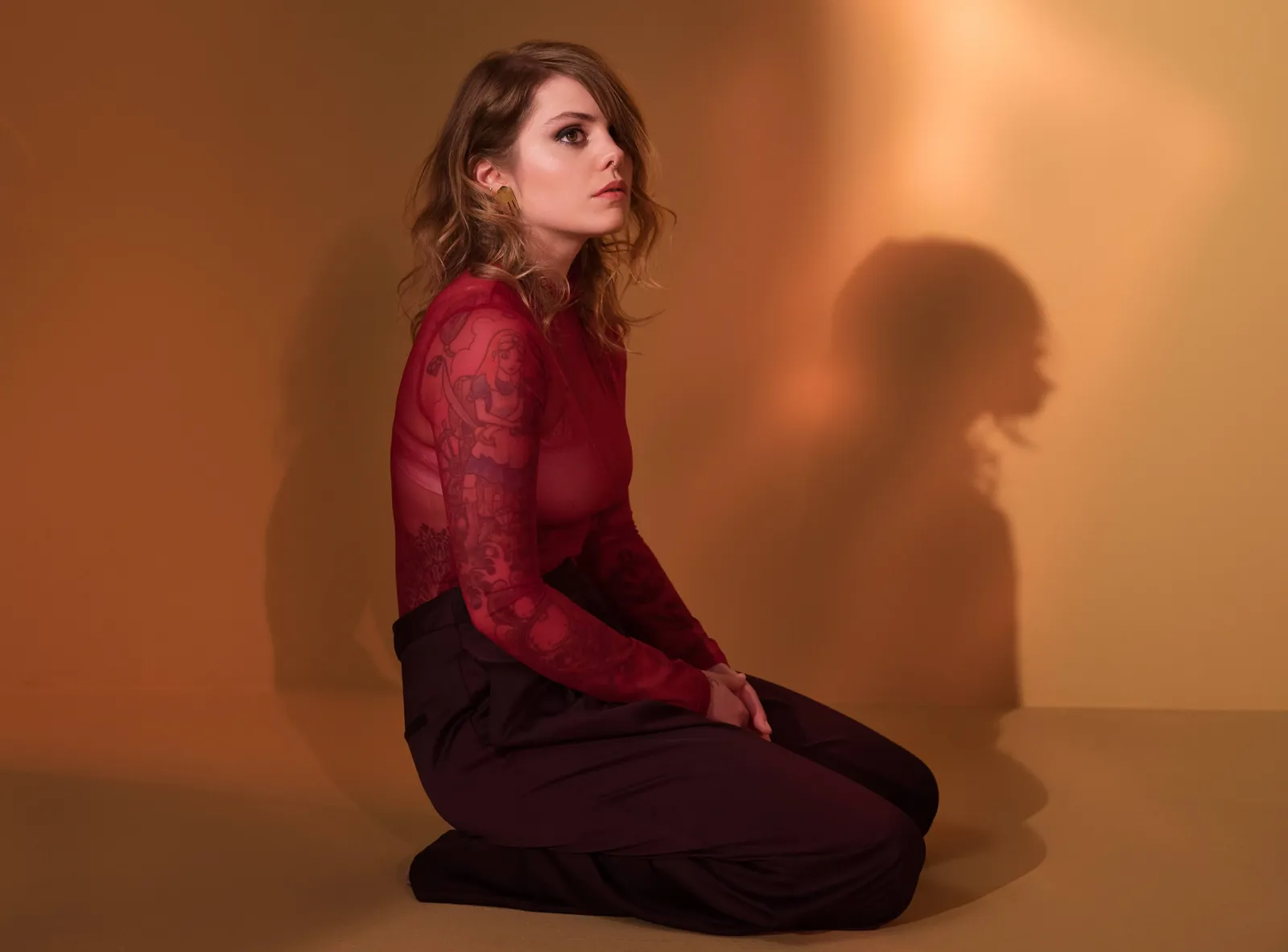 Coeur de Pirate kneels in a sheer red top against an amber wall, her shadow cast beside her