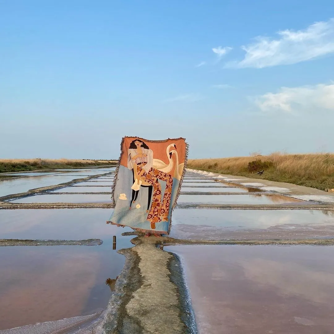 Woven blanket printed with a woman and a swan, held up on a path between flooded salt pans under a blue sky