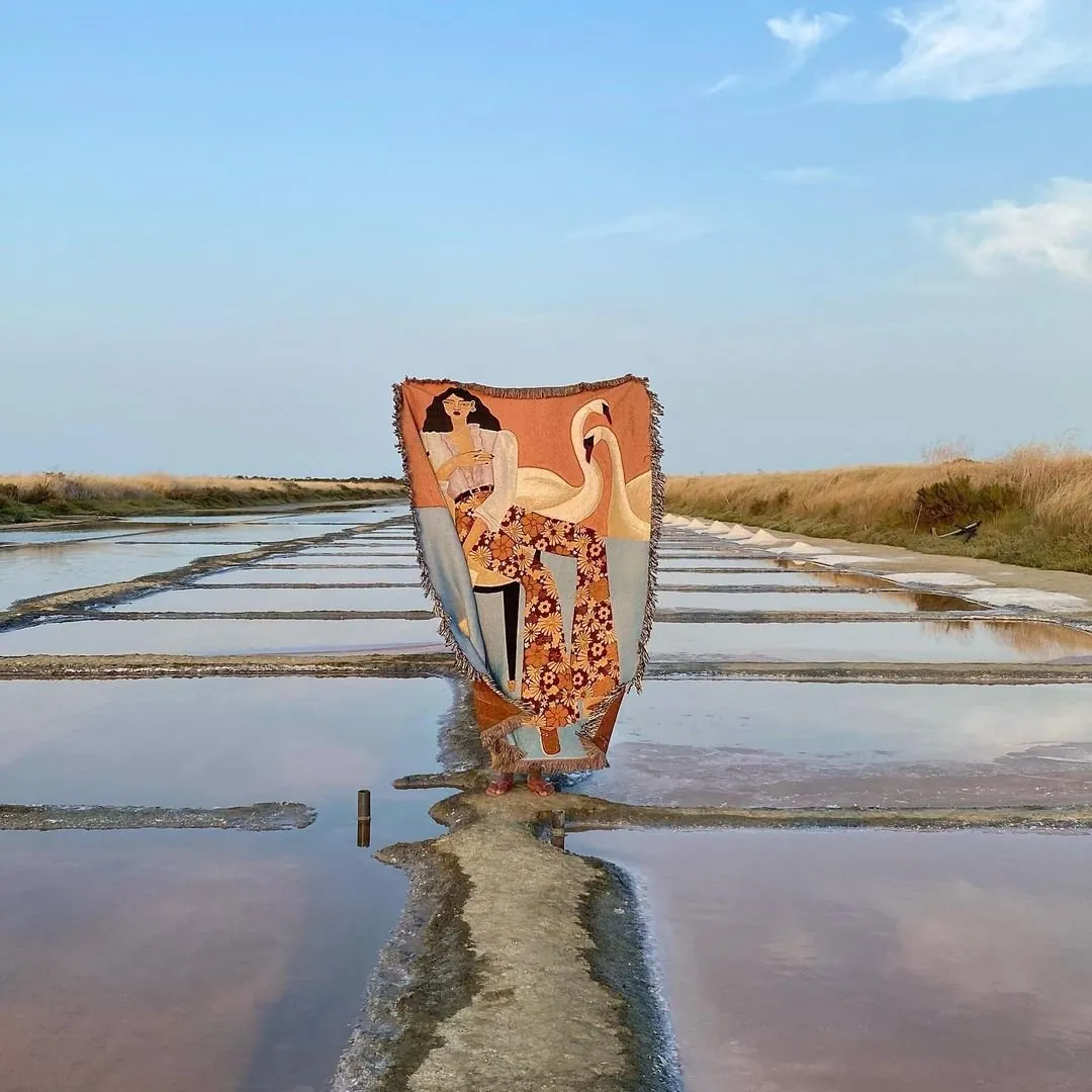 Person hidden behind a woven blanket of a woman with a swan, standing on a narrow path between shallow salt pans
