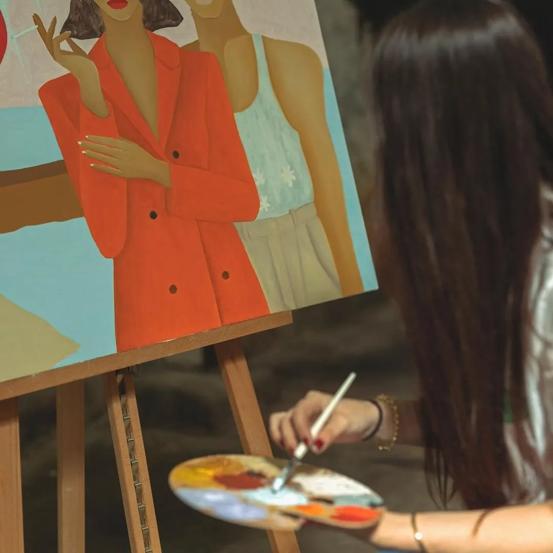 Over-the-shoulder view of Léa Augereau painting a canvas of two women, one in a red blazer holding a cigarette