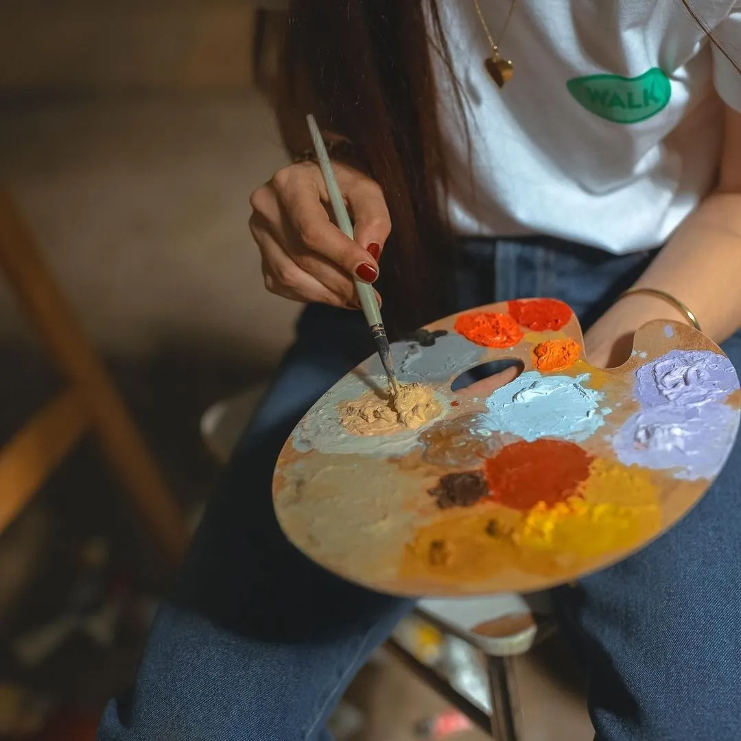 Close-up of a hand with red nails mixing paint on a wooden palette of orange, lilac, blue and yellow