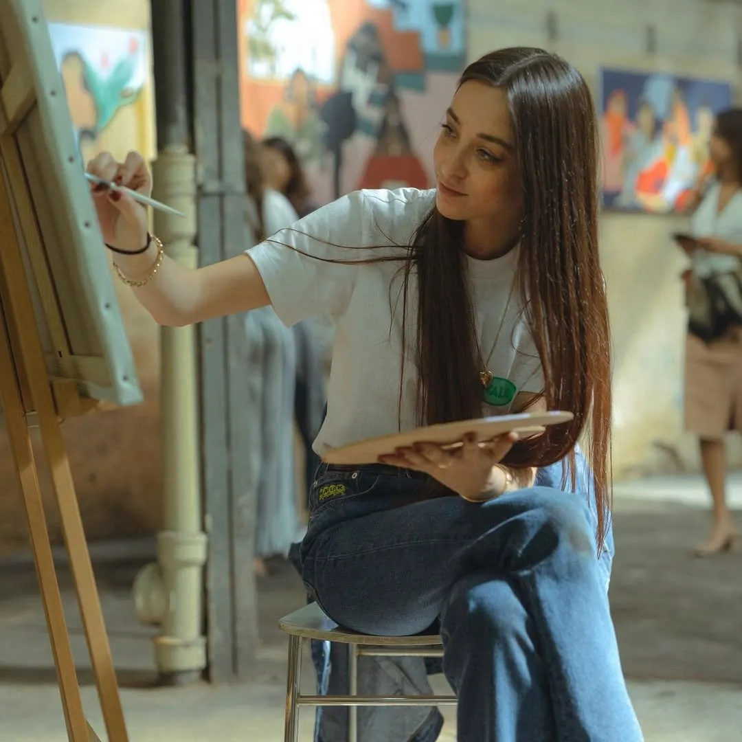 Léa Augereau seated on a stool, brush raised to a canvas on an easel, holding a palette, paintings hanging behind