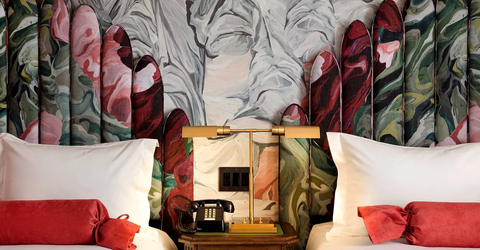 A guest room headboard upholstered in a painted floral fabric, with a brass lamp and a rotary phone