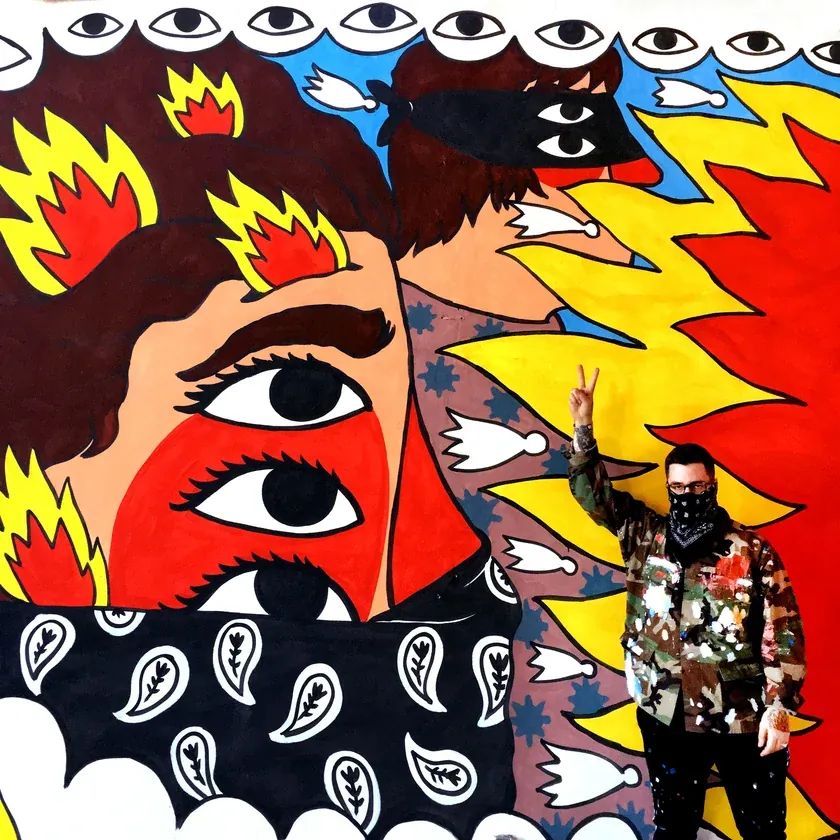 Ricardo Cavolo flashes a peace sign beside a mural of a bandana-masked figure with stacked eyes and flames