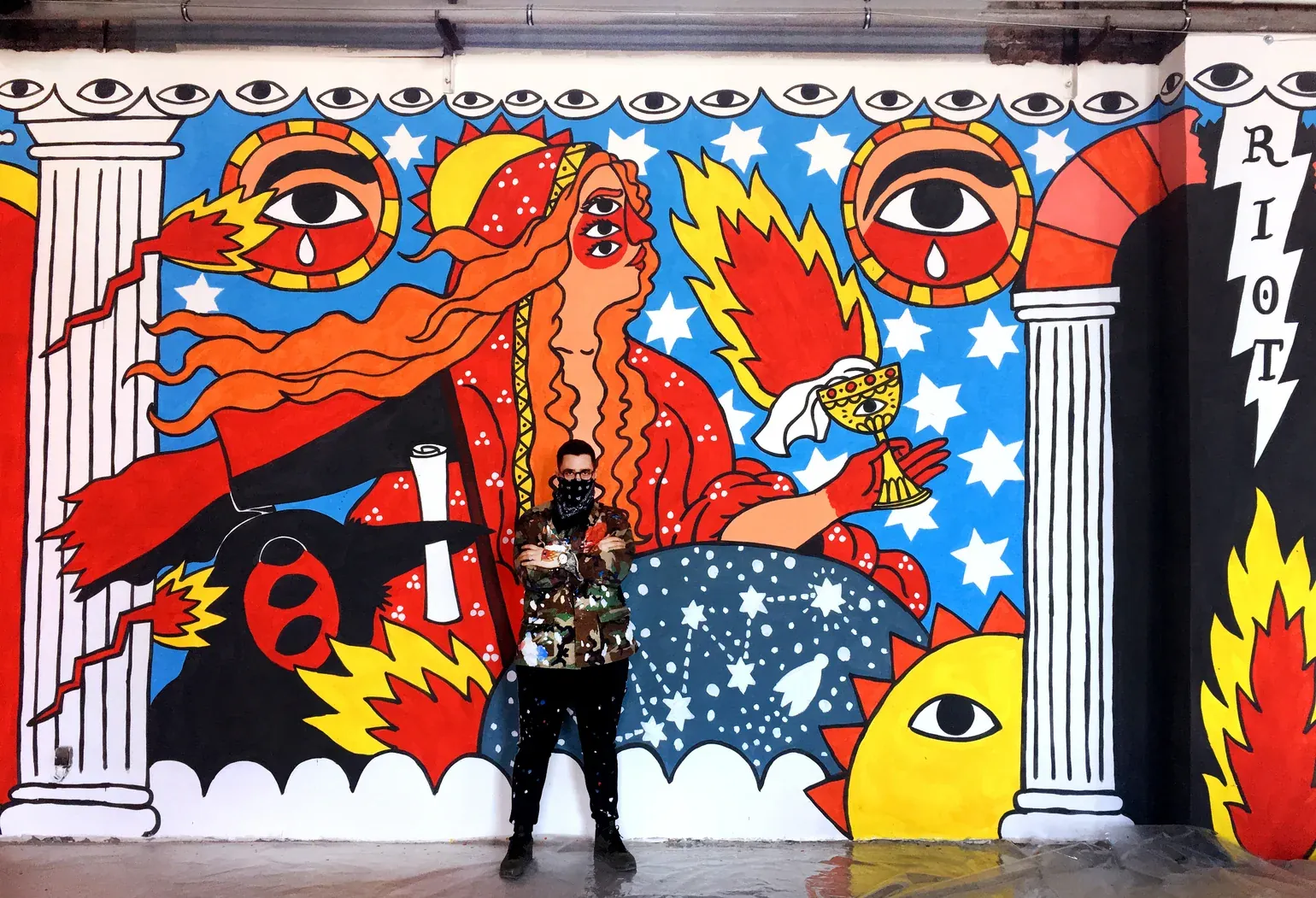 Ricardo Cavolo stands in front of a large mural of a many-eyed woman holding a goblet, with suns, stars and the word RIOT